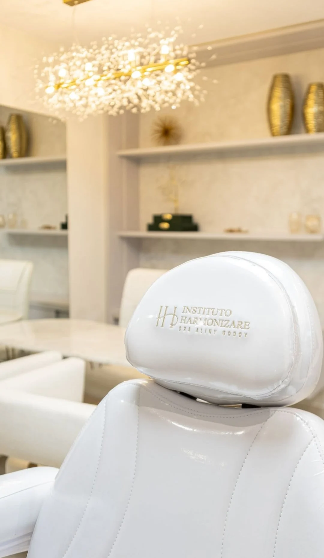 Encosto da cadeira branca de atendimento com o logo do Instituto Harmonizare, Dra Aliny Godoy, bordado; ao fundo, o lustre e as prateleiras do consultório