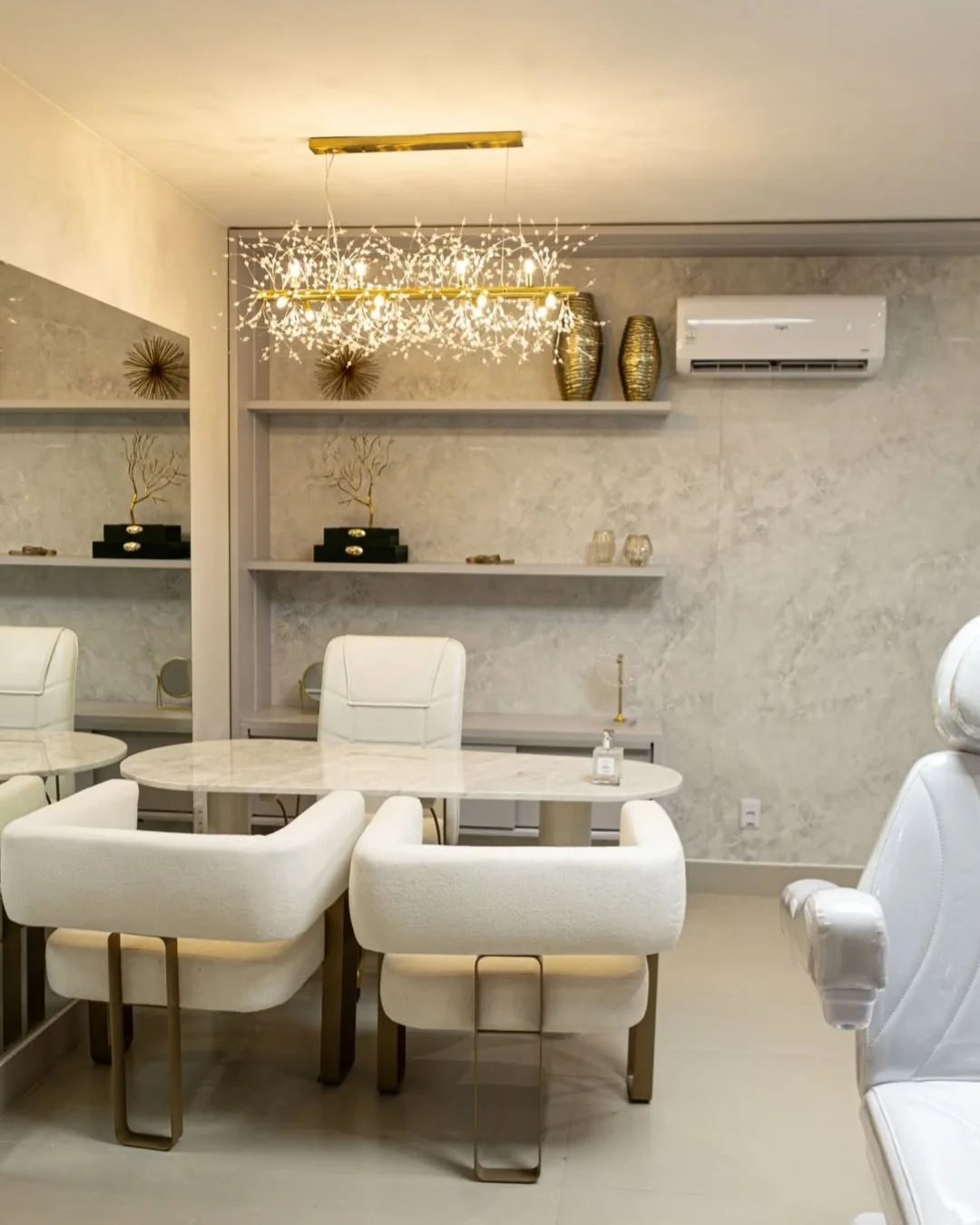 Consultório do Instituto Harmonizare: mesa de mármore com poltronas de bouclé sob um lustre de fios com pontos de luz, prateleiras com vasos dourados e, à direita, a cadeira de atendimento
